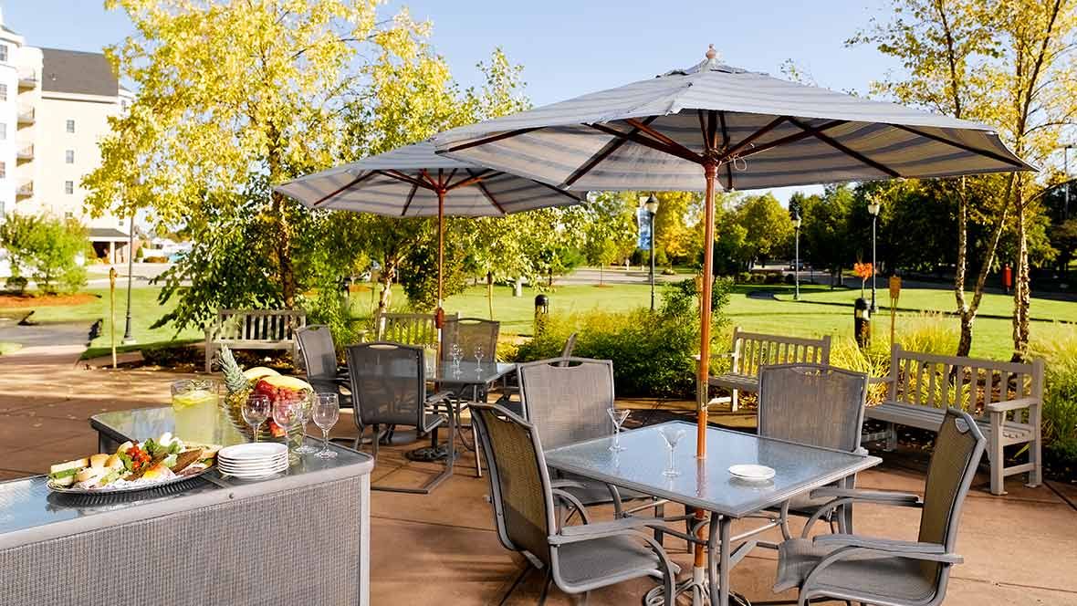 A sunny outdoor patio with tables, chairs, and umbrellas set in a garden setting.