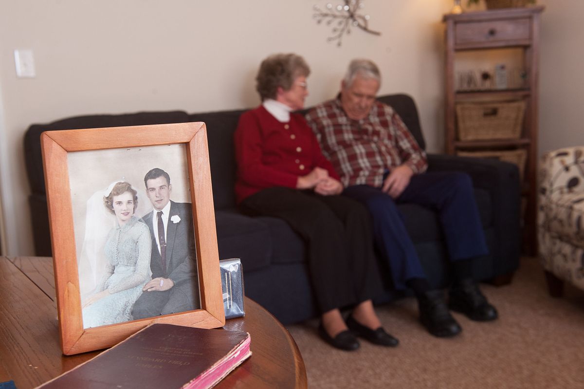 An elderly couple sits on a couch holding hands with an old photograph in the foreground.