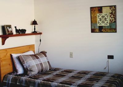 A neatly arranged single bed with checkered bedding, a decorative lamp on a shelf, and framed artwork on the wall.