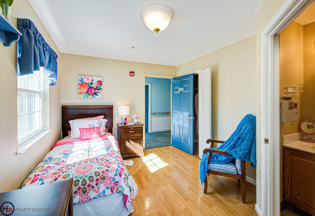 A cozy bedroom with a single bed and colorful bedding, featuring wooden floors, a window, and a chair.