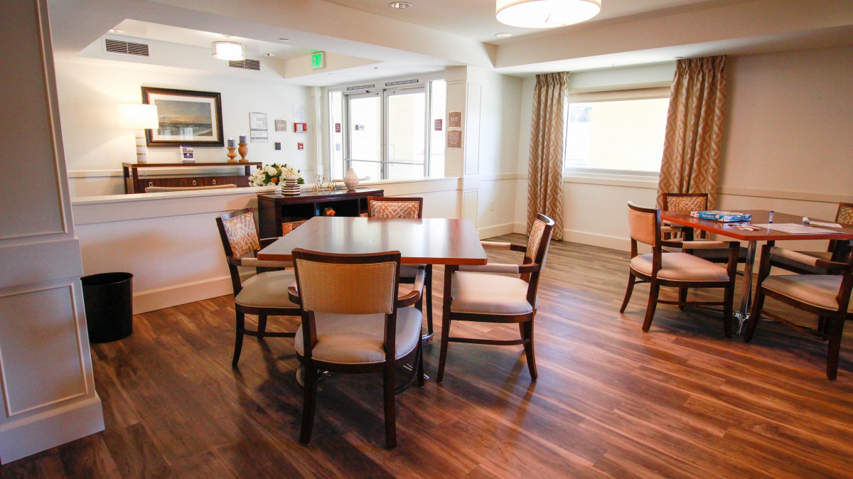 A cozy dining area with tables and chairs set on wooden flooring, illuminated by natural light filtering through large windows.