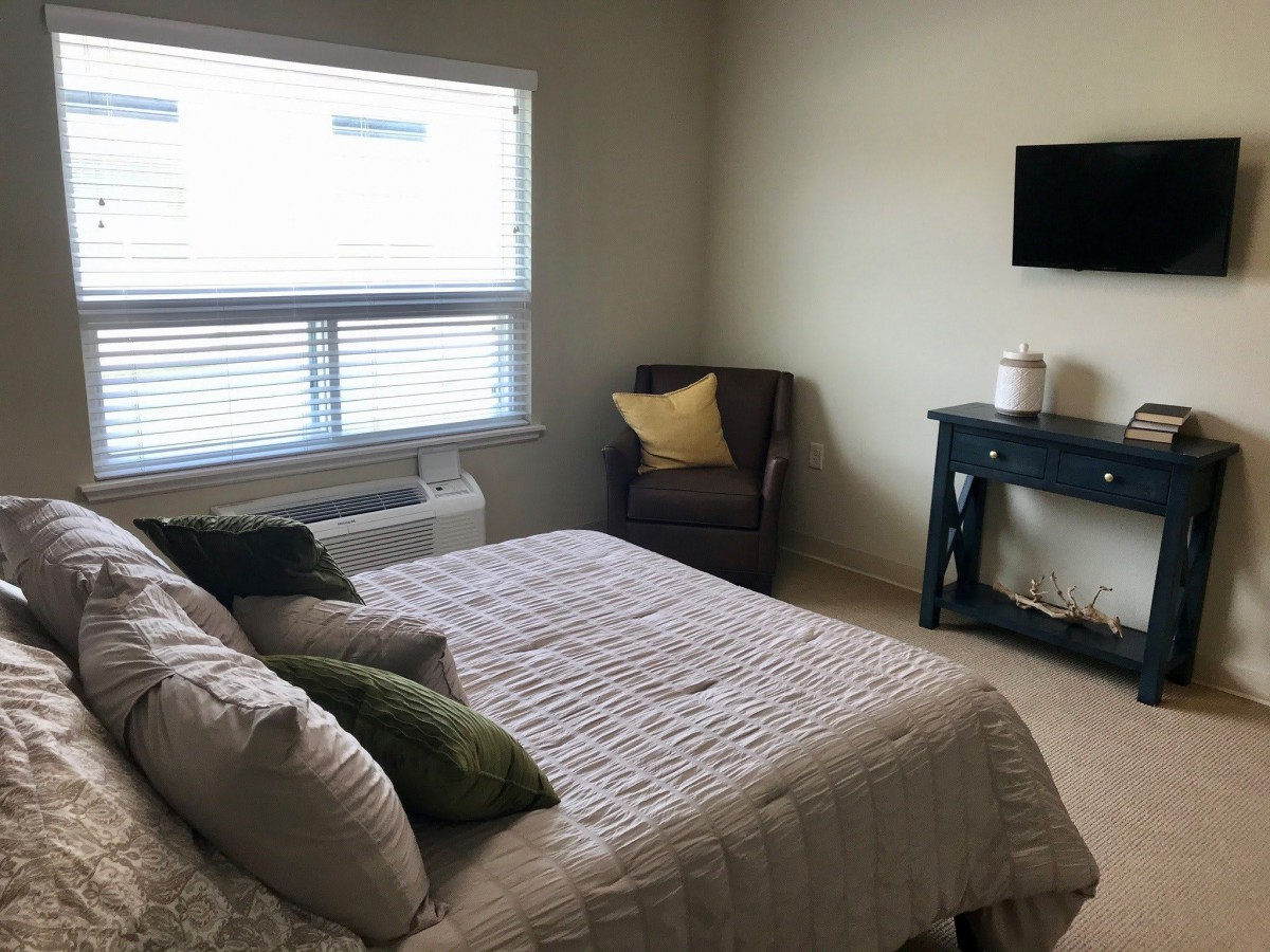 A cozy bedroom featuring a neatly made bed, an armchair with cushions, and a mounted television.