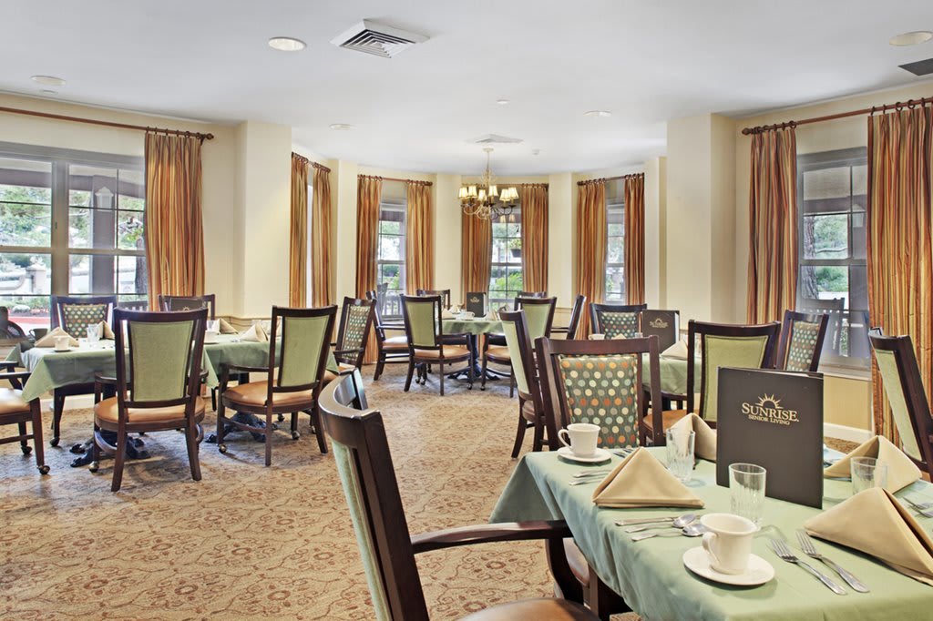 An elegant dining room with neatly arranged tables and chairs, featuring warm lighting and large windows with curtains.