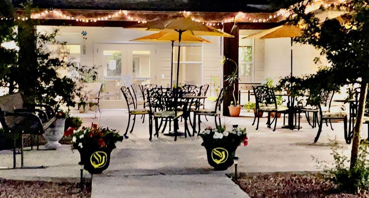 A cozy patio with tables, chairs, and yellow umbrellas, adorned with string lights and surrounded by potted plants.
