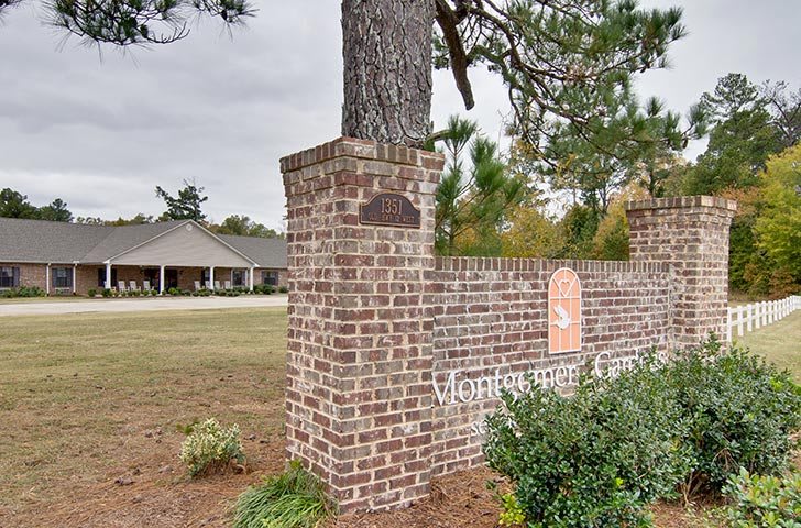 A brick signpost stands at the entrance to a property with a building visible in the background, surrounded by trees and greenery under an overcast sky.