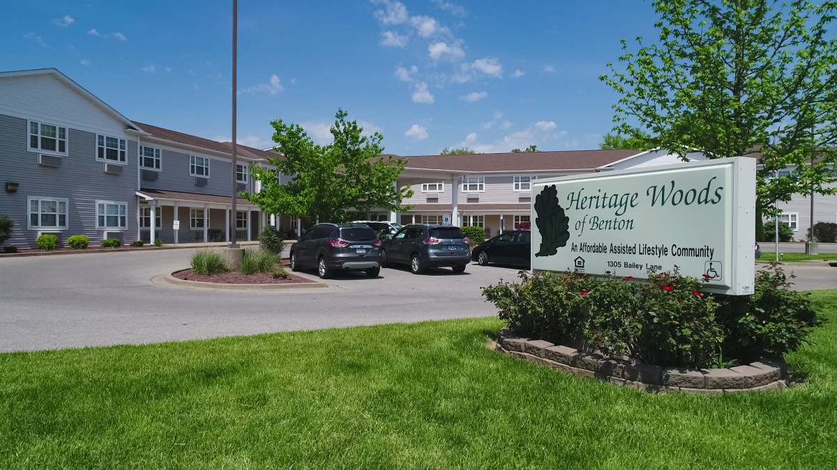 A view of a building with a parking lot, cars, and greenery under a clear blue sky.