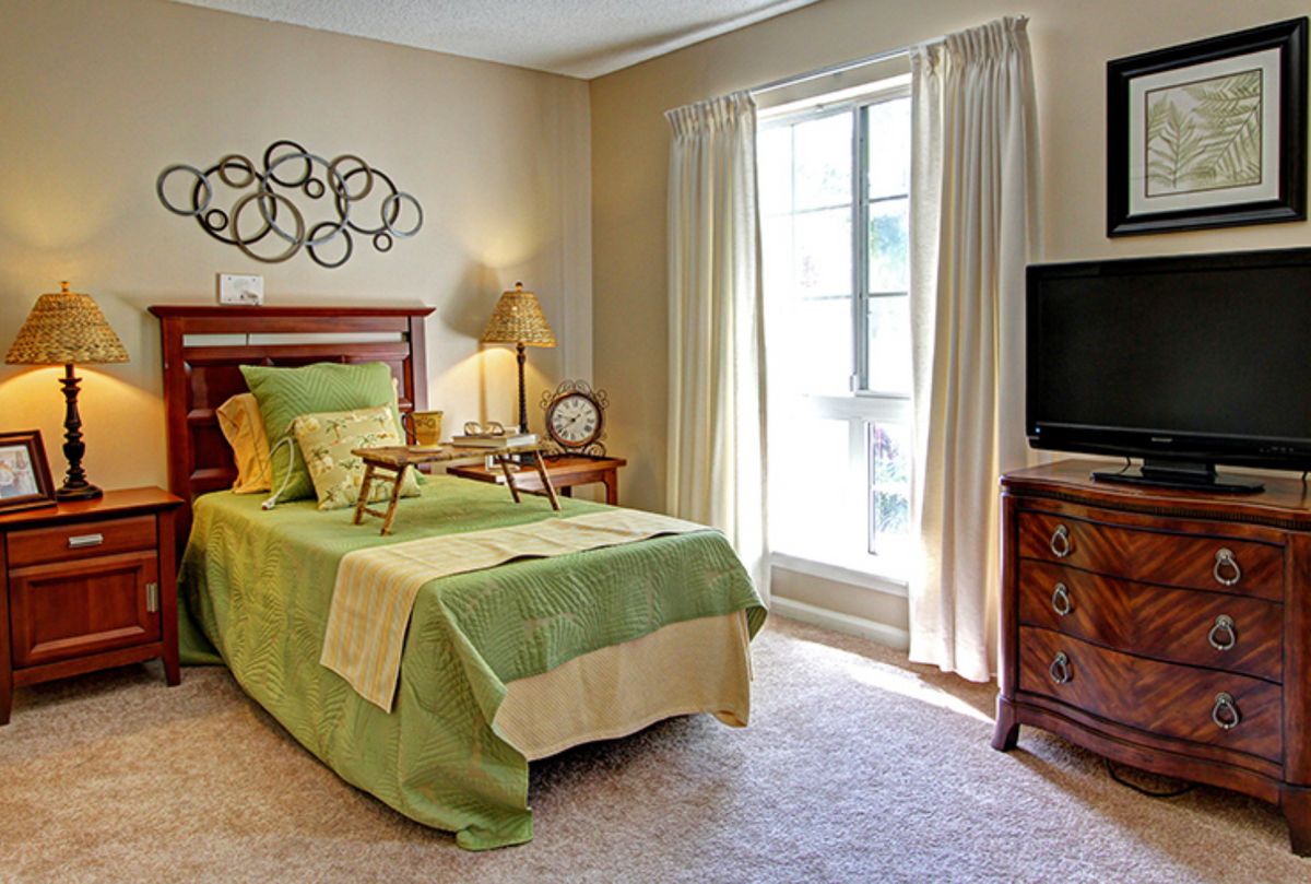 A cozy bedroom with a green-themed bedspread, wooden furniture, and natural light from a large window.
