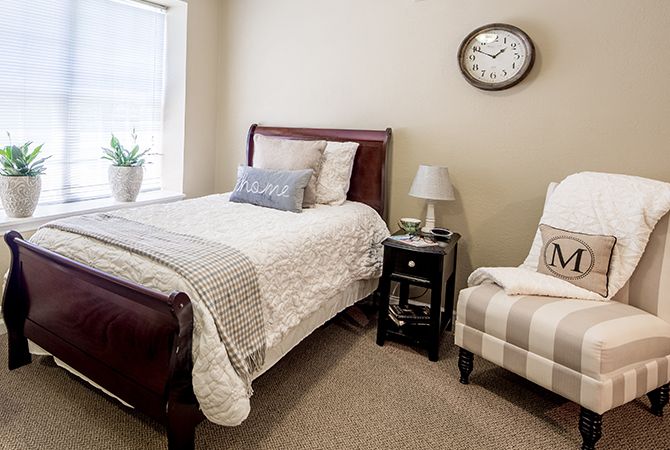 A cozy bedroom with a single bed, nightstand, and chair, featuring neutral tones and minimal decor.