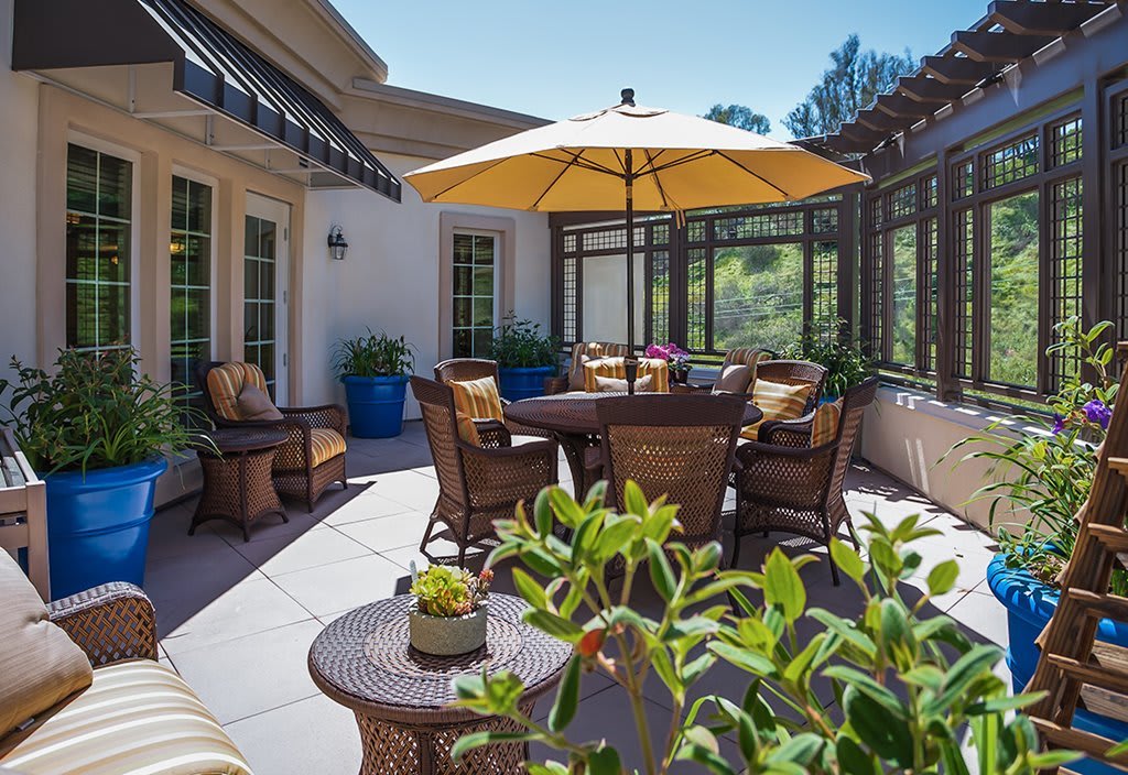 A sunny patio with outdoor furniture featuring a table, chairs, and an umbrella surrounded by potted plants.
