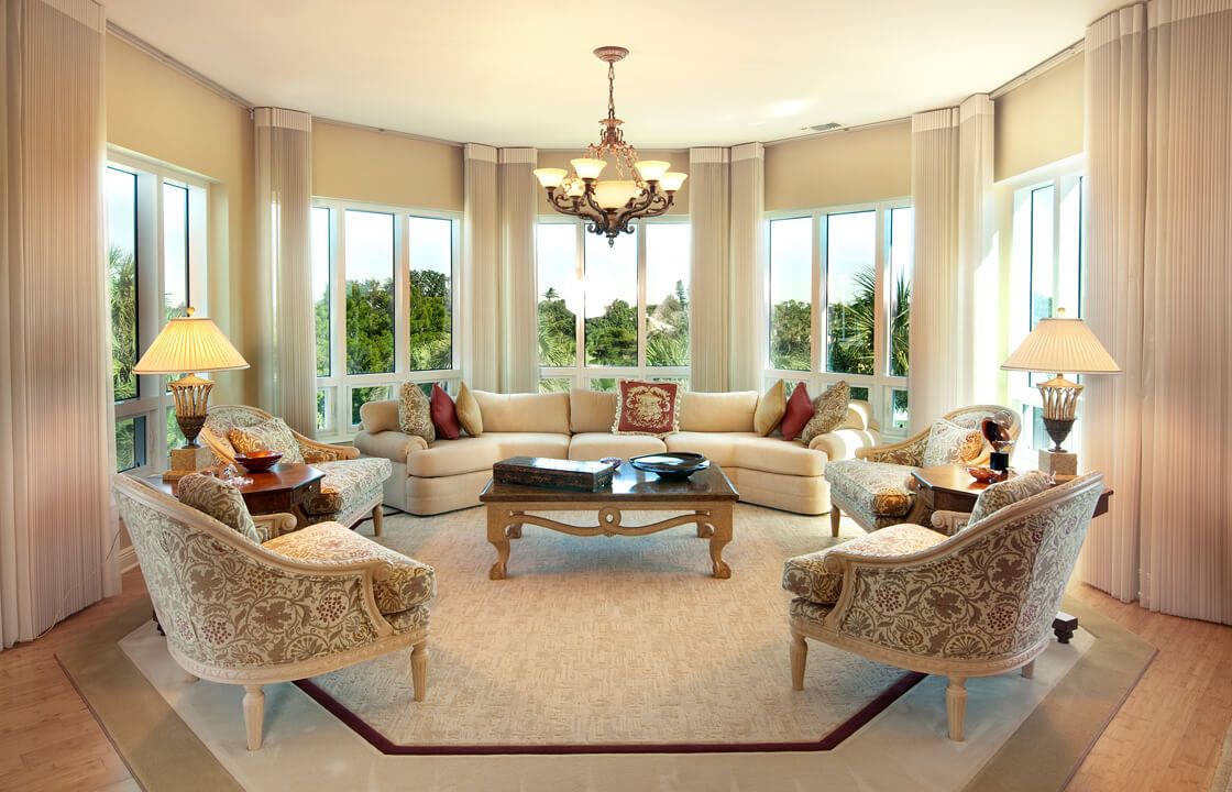 A spacious and elegantly designed living room with a large sectional sofa, a central coffee table, and ample natural light from surrounding windows.