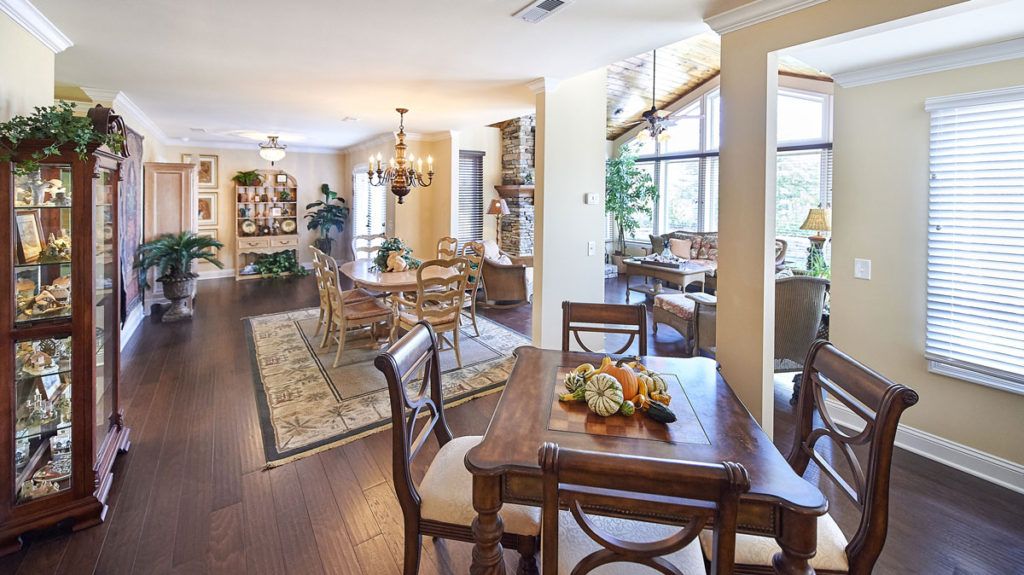 A spacious dining room with elegant wooden furniture, large windows, and a chandelier, featuring plants and decorative elements.