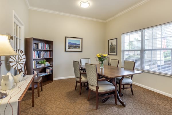 A cozy dining area with a wooden table, chairs, bookshelf, and large window.