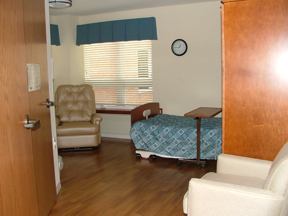 A hospital room with a single bed, armchair, and wooden flooring.