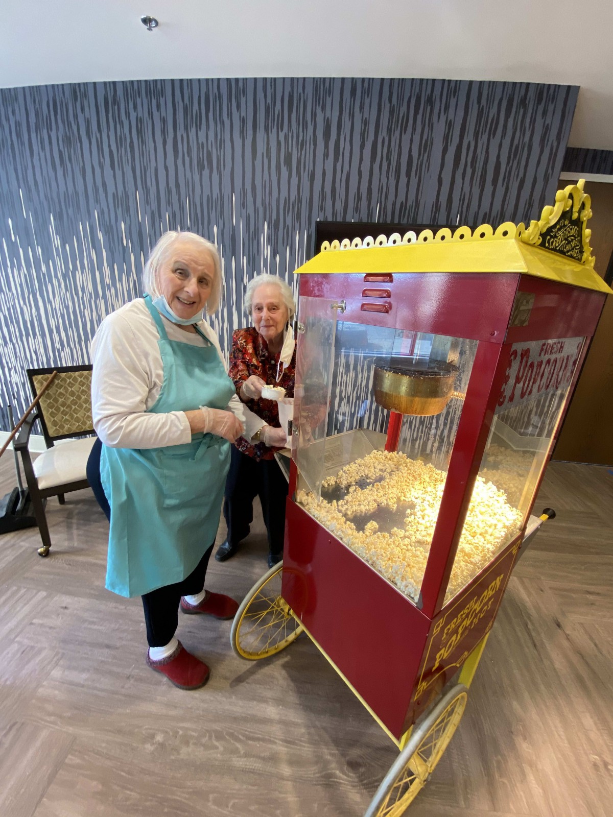 Two elderly women smiling next to a popcorn machine indoors.