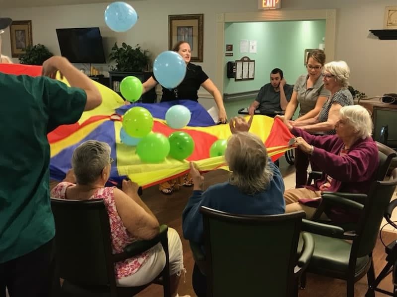 A group of seniors engaging in an indoor activity with a colorful parachute and balloons.