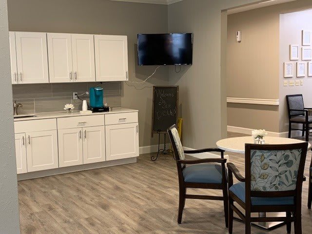 A modern kitchenette area with white cabinets, a small dining table and chairs, and a wall-mounted television.