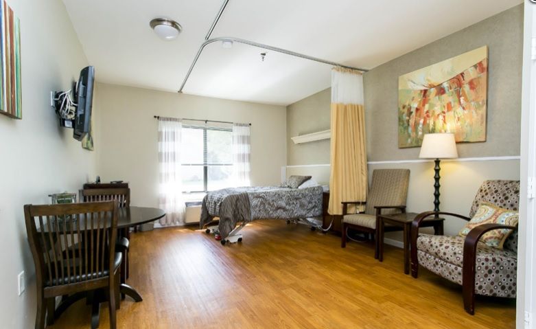 A cozy nursing home bedroom with a single bed, wood flooring, and ample natural light from a large window.