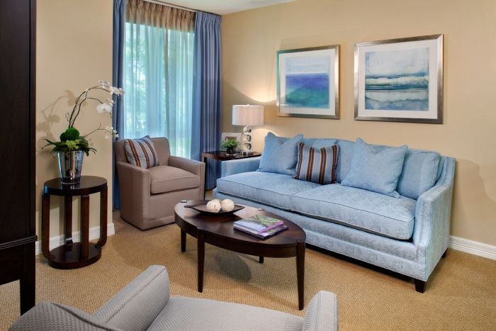 A cozy living room with a blue sofa, armchair, and a wooden coffee table, adorned with artwork and curtains.