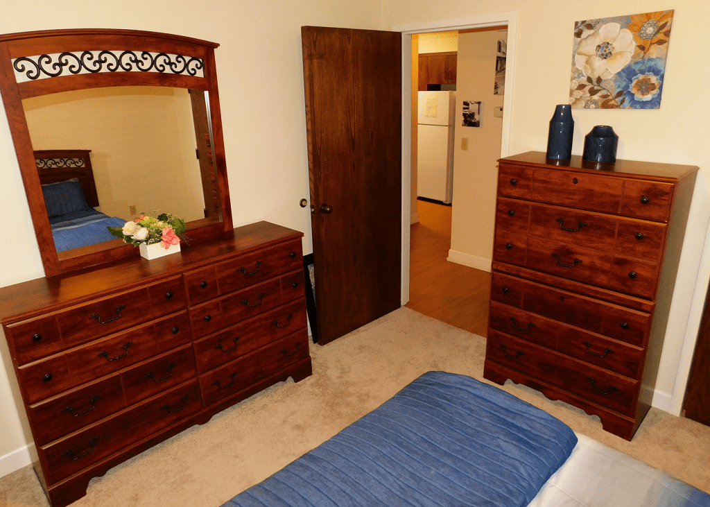 A cozy bedroom with wooden furniture, including a dresser and mirror, leading to a kitchen area.