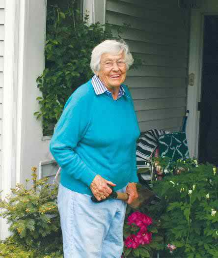 An elderly woman smiling while standing in a garden near a porch.