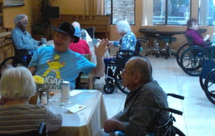 A group of elderly individuals in a nursing home setting, engaged in conversation while seated at a table.