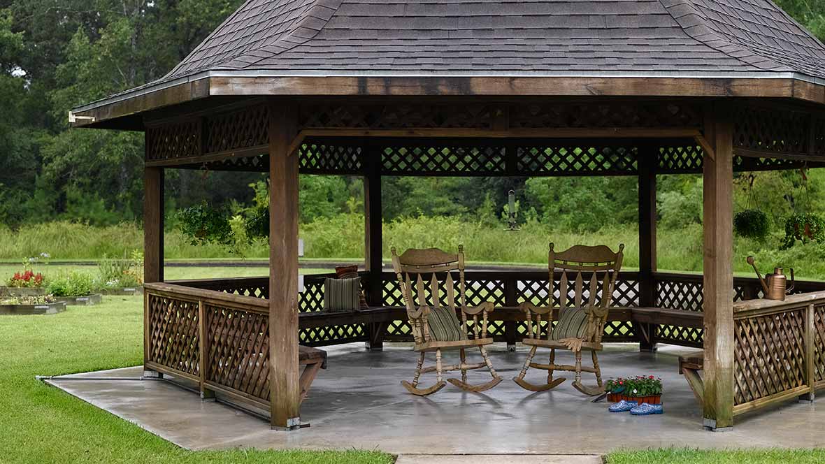 A wooden gazebo with rocking chairs in a garden setting.