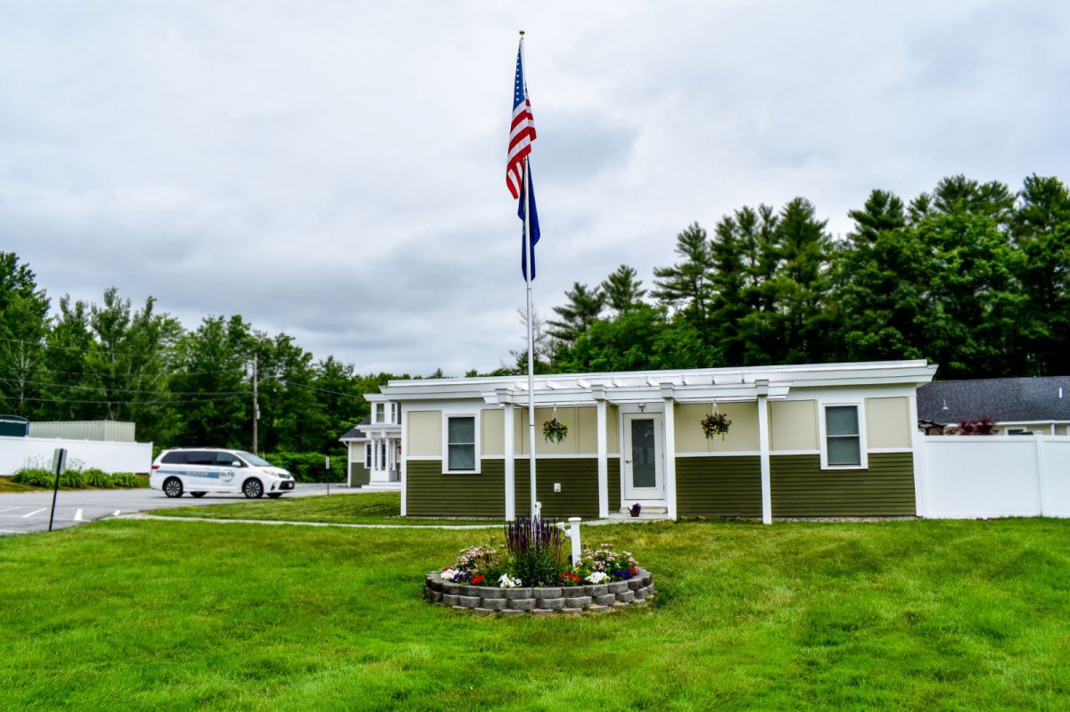A small house with an American flag on a flagpole, surrounded by a well-maintained lawn and trees under a cloudy sky.