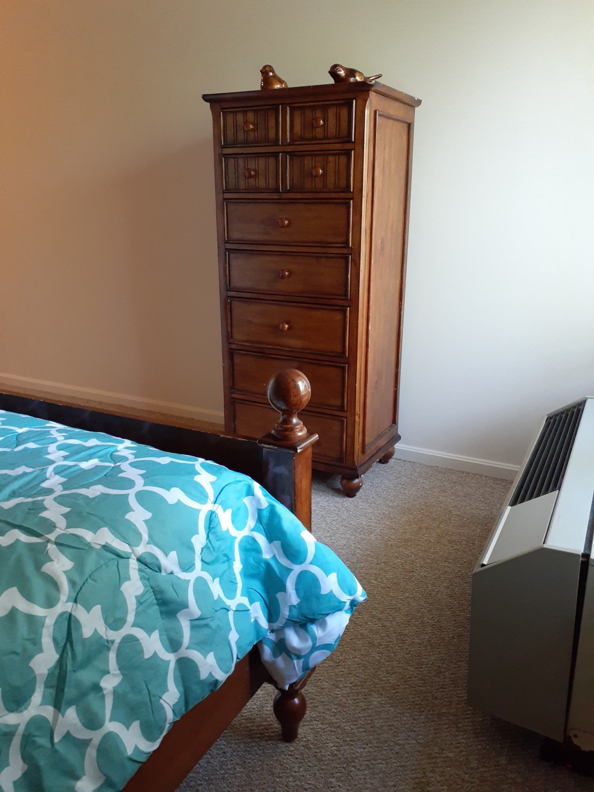 A cozy bedroom featuring a teal quilt, wooden dresser, and a space heater.