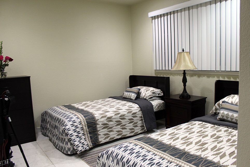 A bedroom with two neatly made twin beds, a bedside table with a lamp, and window blinds.