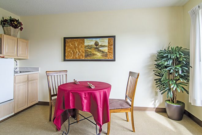 A cozy dining area with a small table covered with a red cloth, surrounded by wooden chairs, and adorned with a potted plant and wall art.
