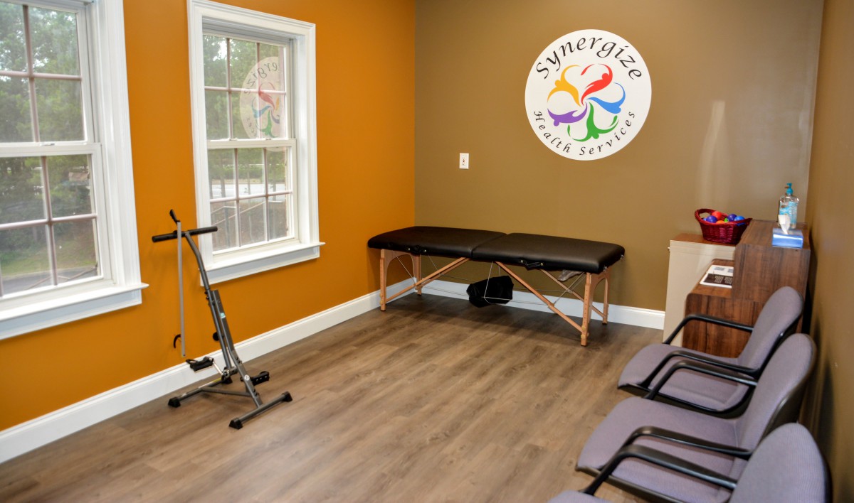 A therapy room with an exercise bike, massage table, and chairs, featuring a colorful logo on the wall.