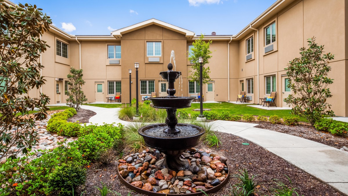 A serene courtyard featuring a decorative fountain surrounded by greenery and a multi-story building.