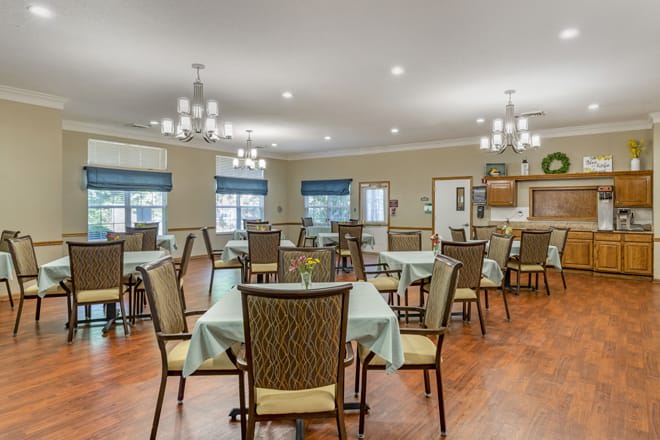A well-lit dining area with neatly arranged tables and chairs, featuring elegant chandeliers and large windows.