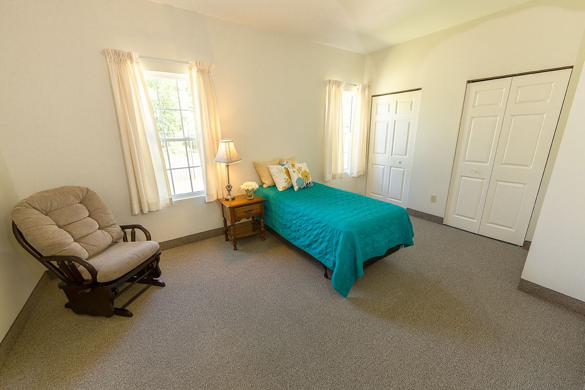 A cozy bedroom with a teal bedspread, large windows, and simple furnishings.
