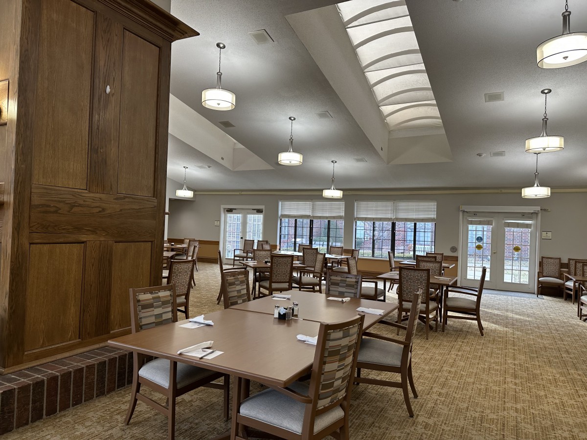 A spacious dining room with neatly arranged tables and chairs, illuminated by ceiling lights and natural light from large windows.