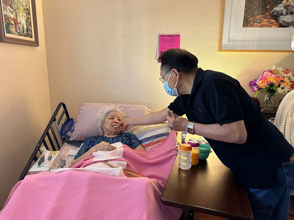 A visitor wearing a mask comforts an elderly patient in a hospital bed.
