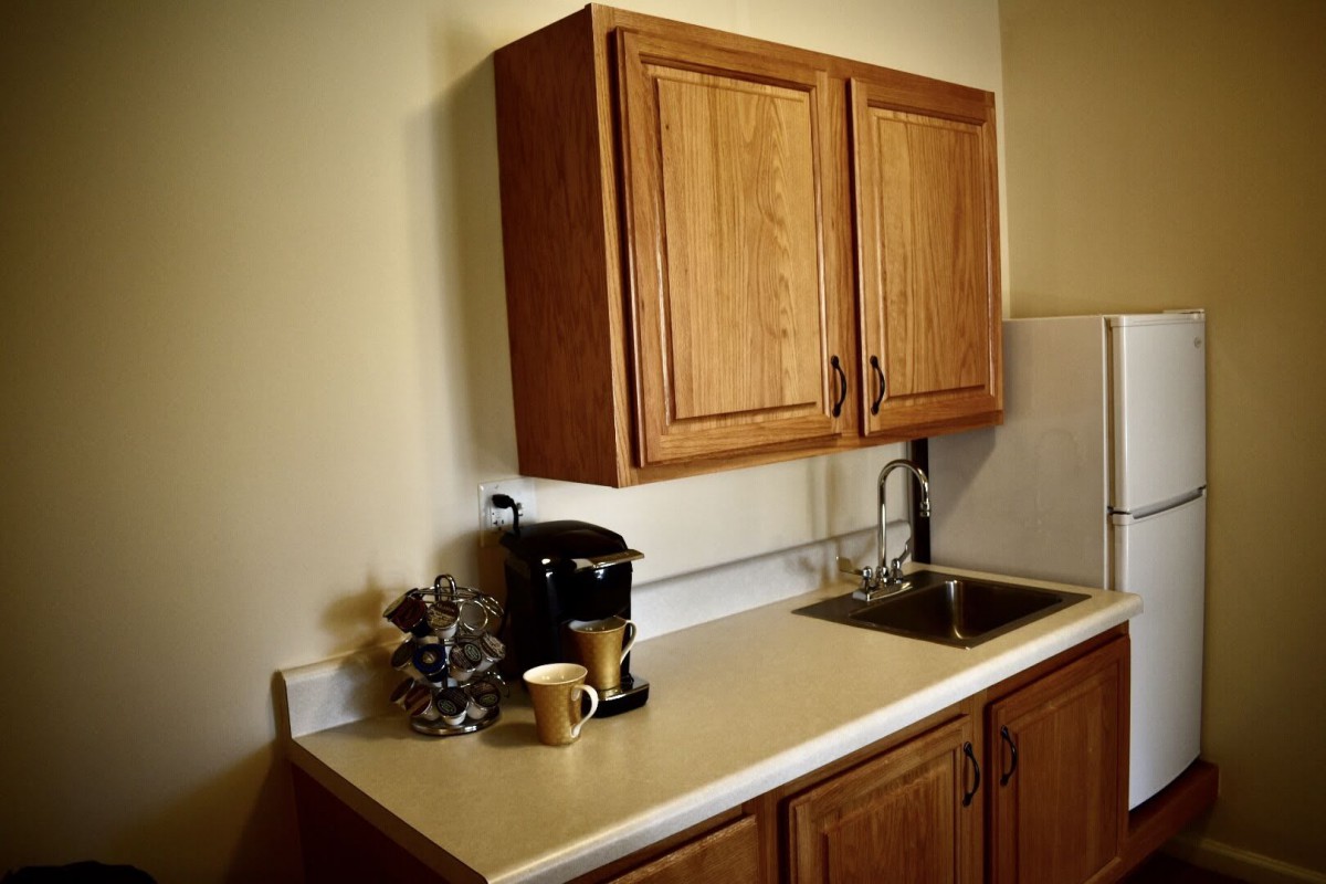 A compact kitchen setup featuring wooden cabinets, a countertop with a coffee maker, mugs, and a small sink.