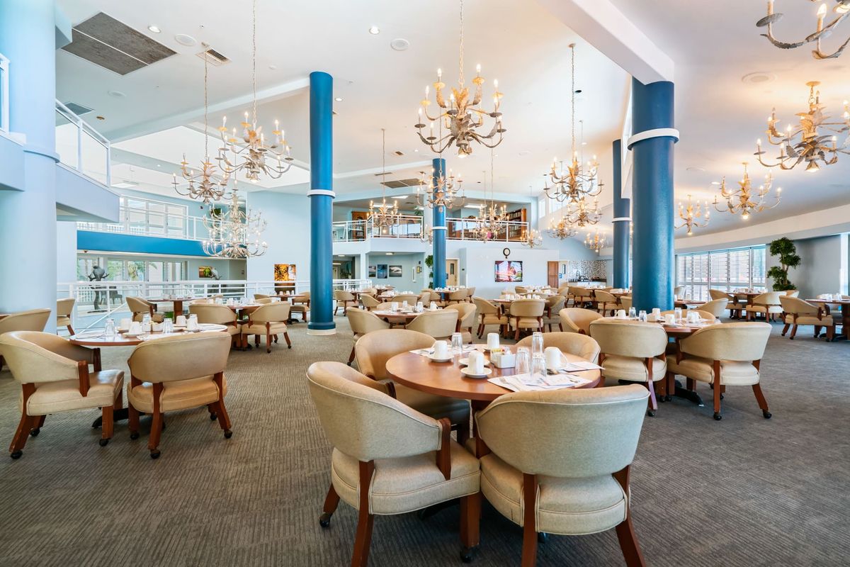 A spacious and elegantly designed restaurant with chandeliers hanging from the ceiling, set up with round tables and plush chairs.