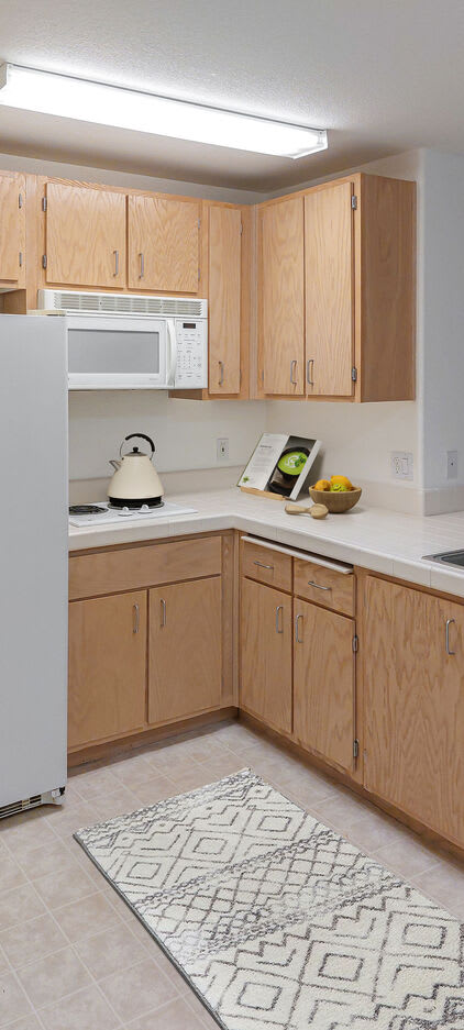 A modern kitchen with wooden cabinets, a white countertop, and various kitchen items including a microwave, kettle, and fruit bowl.