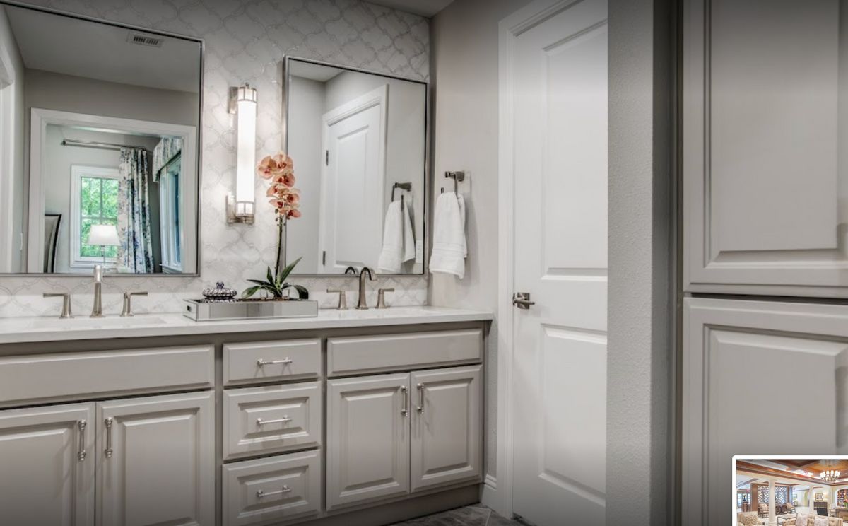 A modern bathroom with a large mirror, sink, and white cabinetry, featuring a decorative plant and neatly hung towels.