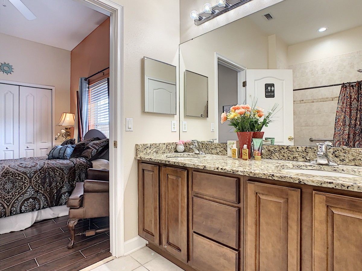 A bathroom with a granite countertop and wooden vanity, featuring flowers in a vase, with a glimpse into an adjacent bedroom.