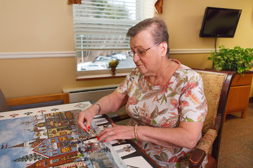 An elderly woman enjoys working on a puzzle at a well-lit table.