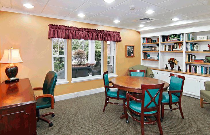 A cozy room with a round table surrounded by chairs, a large window with curtains, and bookshelves filled with various items.