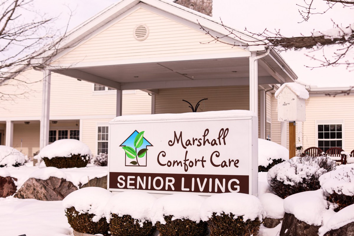 A snow-covered sign for Marshall Comfort Care Senior Living in front of a building.