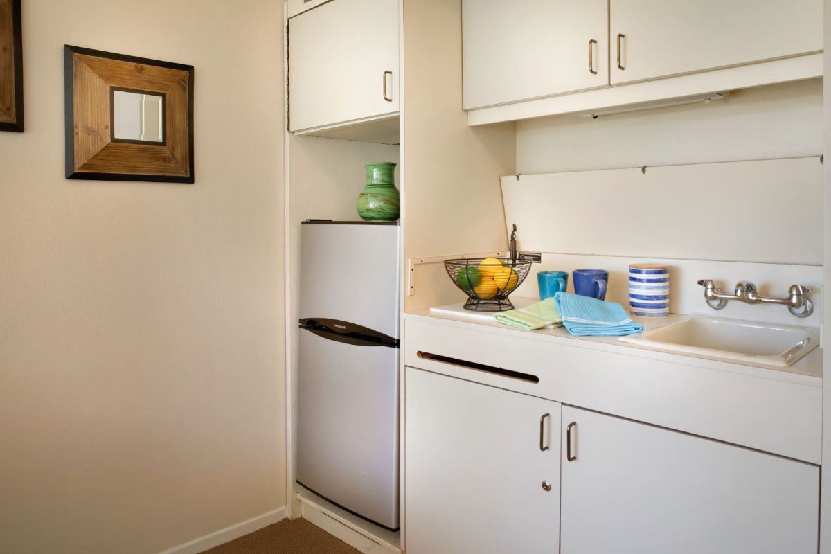 A compact kitchen corner with white cabinets, a small refrigerator, and a tidy countertop featuring a fruit bowl and some kitchen items.