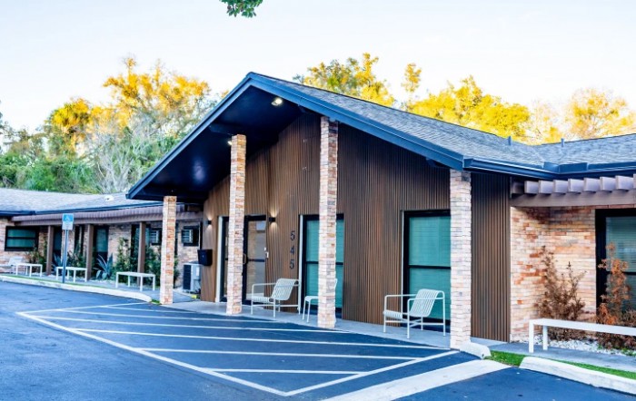 A small building with a sloped roof, surrounded by trees and a parking lot.