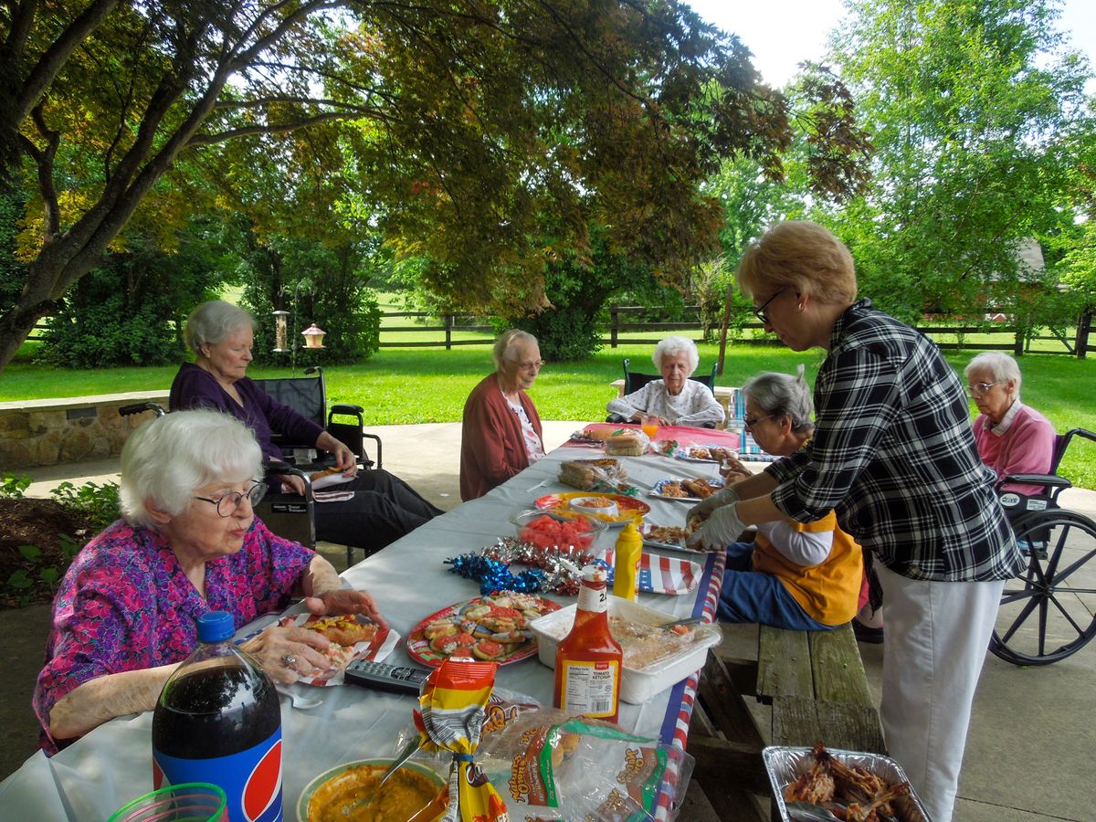 A group of people enjoying a picnic at a park table, surrounded by trees and an assortment of food and drinks.