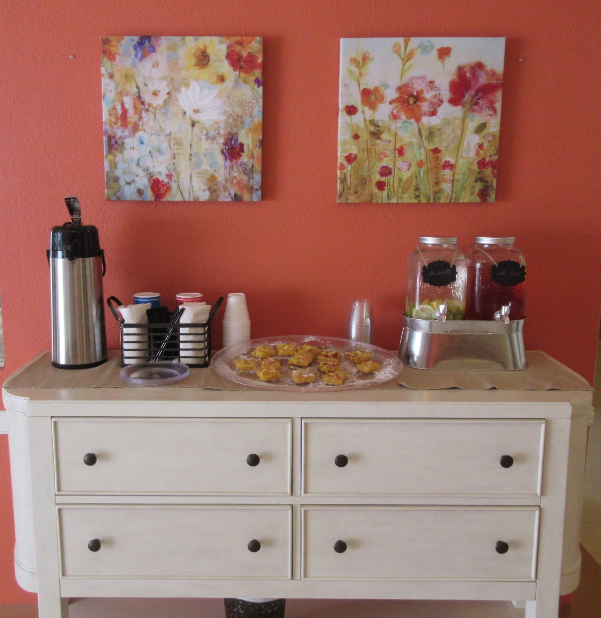 A cozy refreshment station with coffee, cups, and pastries displayed on a white buffet table against a warm orange wall adorned with floral artwork.