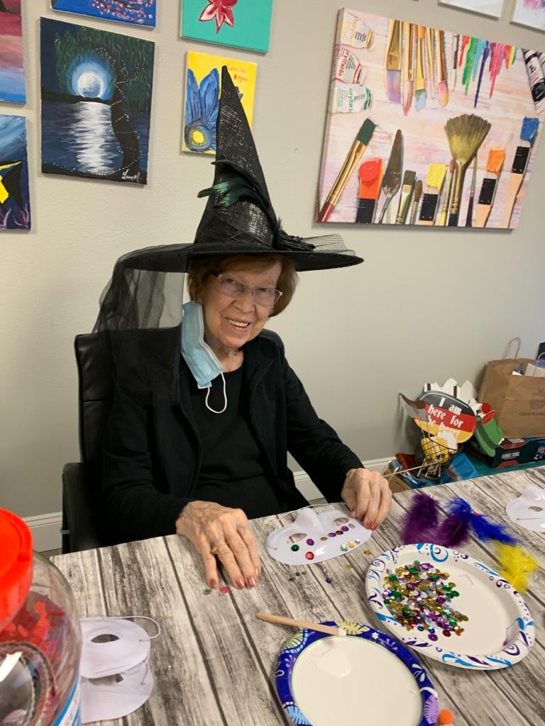 A person wearing a witch hat sits at a table filled with art supplies, engaging in Halloween-themed crafts.
