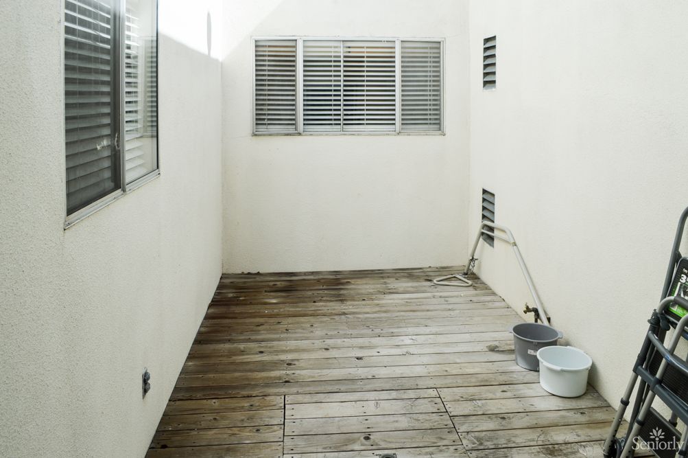 A minimalist outdoor patio with wooden flooring and white walls, featuring window shutters.